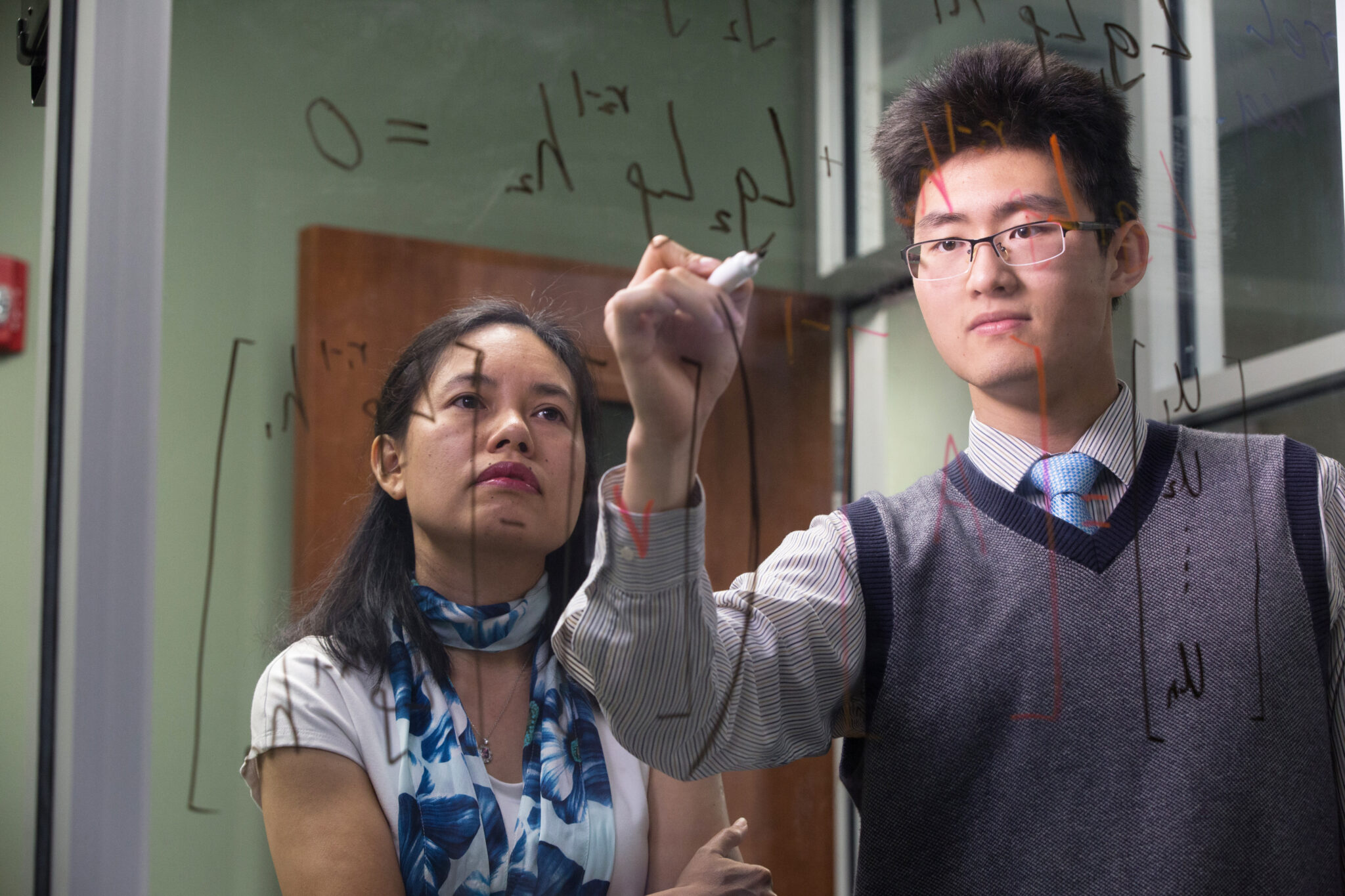 Student working on equation on a dry erase board with a professor helping him