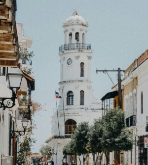 White clock tower of the Palacio Consistorial in Santo Domingo, Dominican Republic, lining a quiet colonial street with trees and historic buildings.