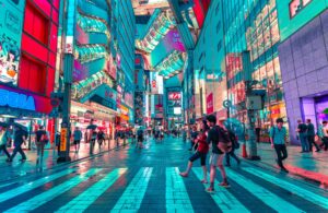 Pedestrians walk along a rain-slicked street in Akihabara, Tokyo at night, surrounded by towering neon-lit buildings, colorful storefronts, and glowing signage in red, teal, and pink.