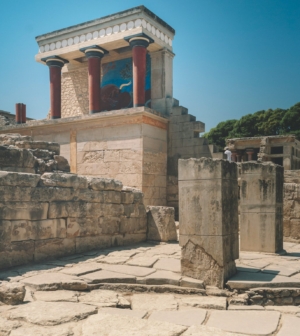 The partially reconstructed North Entrance of the Minoan Palace of Knossos, featuring distinctive red columns and vivid frescoes atop ancient stone ruins under a clear blue sky, Crete, Greece.