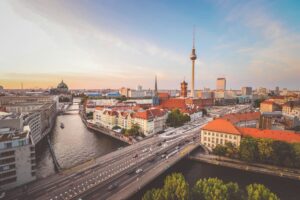 Aerial view of Berlin at golden hour, with the iconic Fernsehturm television tower rising above a cityscape of red-roofed buildings, bridges, and the winding Spree River, Germany.