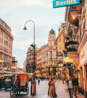 Pedestrians walk along a bustling European shopping street lined with ornate historic buildings, glowing storefronts, and a Berlitz language school sign, Vienna, Austria.