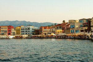 The colorful waterfront of Chania's Old Venetian Harbor glowing with warm light at dusk, with crowds strolling along the promenade backed by pastel buildings and distant mountains, Crete, Greece.